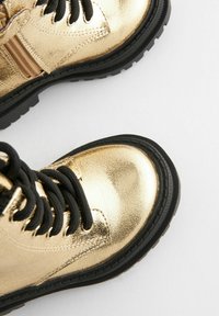 Metallisch goldene Stiefeletten mit schwarzen Schnürsenkeln, strukturiertem Obermaterial, runder Fußspitze und dicker schwarzer Gummisohle, versehen mit akzentuierten Nähten.