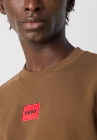 Artimų planų nuotrauka, kurioje matyti žmogus, vilkintis rudą apvalios iškirptės megztinį su raudonu stačiakampiu ženkleliu, pažymėtu žodžiu "HUGO" ant krūtinės.