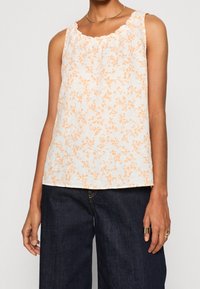 Haut blanc sans manches avec imprimé floral orange, encolure à volants, tissu léger et coupe ample, assorti à un pantalon en denim foncé.