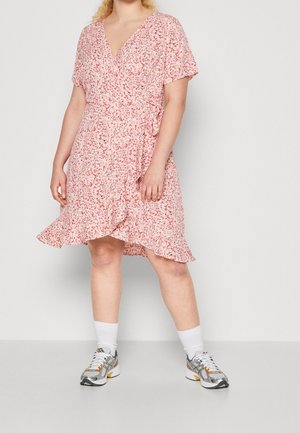 Vrouw die een roze bloemenwikkeljurk met ruches draagt, gecombineerd met witte sokken en zilveren sneakers, staand tegen een effen achtergrond.