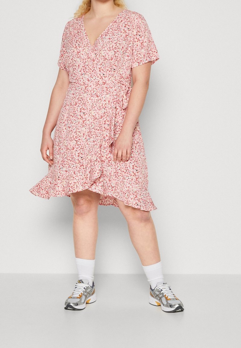 Femme portant une robe portefeuille fleurie rose avec des volants, associée à des chaussettes blanches montantes et des baskets argentées, debout devant un fond uni.