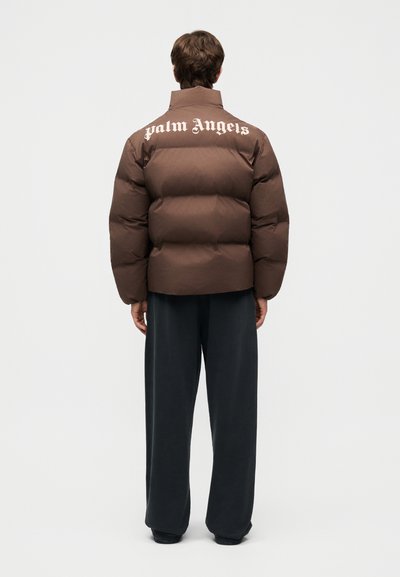 Person trägt eine braune Steppjacke mit dem "Palm Angels"-Schriftzug auf dem Rücken und lockere schwarze Hosen, steht mit dem Rücken zum Betrachter vor weißem Hintergrund.