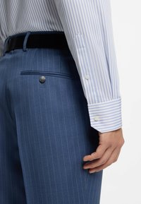 Pantaloni blu a righe sottili con taglio slim, dotati di tasca posteriore con bottone e cintura nera. Polsini di camicia elegante azzurro a righe visibili.