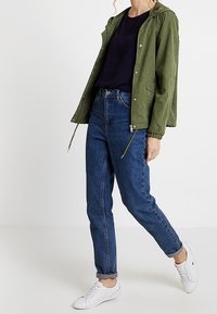Giacca leggera verde oliva con cappuccio, abbinata a una maglia blu e jeans a vita alta blu. Accentuato da sneakers bianche.
