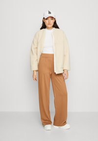 Veste zippée crème, floue, portée sur un t-shirt blanc, associée à un pantalon large à rayures verticales brunes et des baskets blanches, complétée par une casquette blanche.