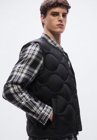 Μαύρο μπουφάν με επένδυση και υφασμένους κυκλικούς μοτίβους που φοριέται πάνω από γκρι και μαύρο καρό πουκάμισο με κουμπιά. Διαθέτει φερμουάρ μπροστά και πλαϊνές τσέπες.