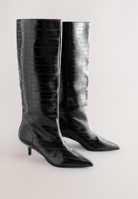 Bottes noires brillantes montant jusqu'au genou, avec des bouts pointus et de petits talons kitten, texturées d'un motif crocodile subtil, reposant sur une surface claire.