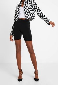 Zwarte biker shorts gecombineerd met een zwart-wit geruite cropped jas. Het model draagt zwarte hakken met bandjes.