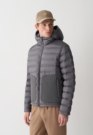 Uomo che indossa una giacca imbottita grigia con cappuccio, grandi tasche frontali, pantaloni beige e un cappello di velluto a coste beige, in piedi contro uno sfondo semplice.