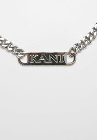 Ezüst színű fém lánc nyaklánc, amelyen egy négyszögletes címke található, melyen vastag betűkkel "KANI" van gravírozva. Sima felület és fényes bevonat jellemzi.