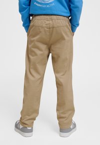 Oseba v bež hlačah z elastičnim pasom in sivimi supergami, roke v žepih, modra jopica z napisom "Everyday Active".