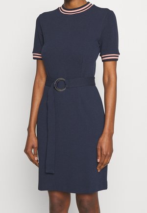 Femme portant une robe bleu marine à manches courtes avec des rayures roses sur le col et les manches, et une ceinture assortie avec une boucle ronde.