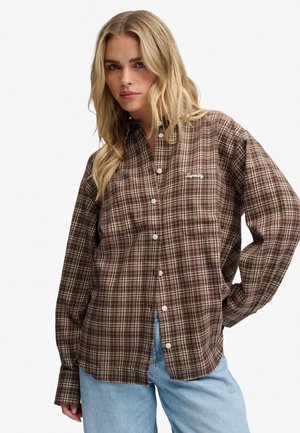 Kobieta w brązowej, kraciastej koszuli zapinanej na guziki z logo "Superdry" i jasnoniebieskich dżinsach, pozująca z jedną ręką na biodrze.