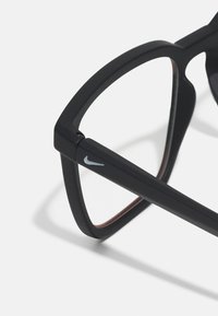 Mustat silmälasit, joissa on matta pinta, neliskulmaiset kehykset ja pieni harmaa Nike-logo sivussa. Ruskeita yksityiskohtia sisäpuolella.