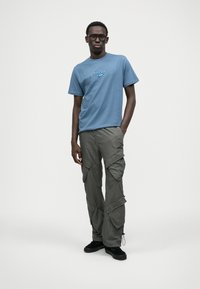 Homem a usar uma t-shirt gráfica azul, calças cargo cinzas, sapatos pretos e óculos está de pé com uma mão no bolso, em frente a um fundo simples.