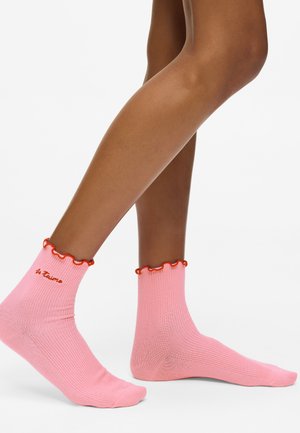 Beine, die pinke gerippte Socken mit roten Rüschensäumen und der Stickerei "Je t'aime" auf einer Seite tragen, vor einem weißen Hintergrund.