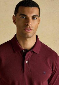 Jeune homme aux cheveux courts et bouclés portant un polo bordeaux avec un petit logo brodé sur la poitrine, debout devant un fond beige.