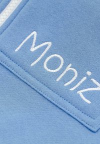 Lichtblauwe stof met een textuur en een geborduurd wit logo "Moniz" dat diagonaal is gepositioneerd, met een soepele steek en een subtiele glans.