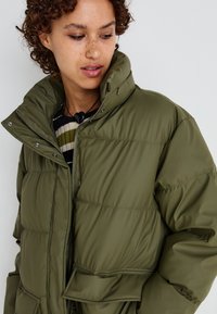 Veste matelassée vert olive avec un col haut, dotée d'une fermeture éclair à l'avant, de boutons-pression et de grandes poches avant. Matériau texturé et léger.