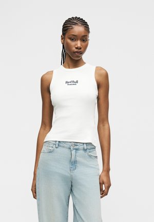 Femeie cu părul împletit, purtând un top alb fără mâneci Red Bull Racing și blugi albastru deschis, stând într-un decor simplu.