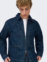 Denimjakke i mørk blå med et strukturert design, med to frontlommer og glidelåslukking. Sømmer detaljer er synlige over hele jakken.