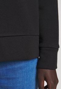 Zwarte sweatshirt met geribde boorden, voorzien van een zachte textuur. Gecombineerd met blauwe denim die een lichte vervaging bij de zoom vertoont.