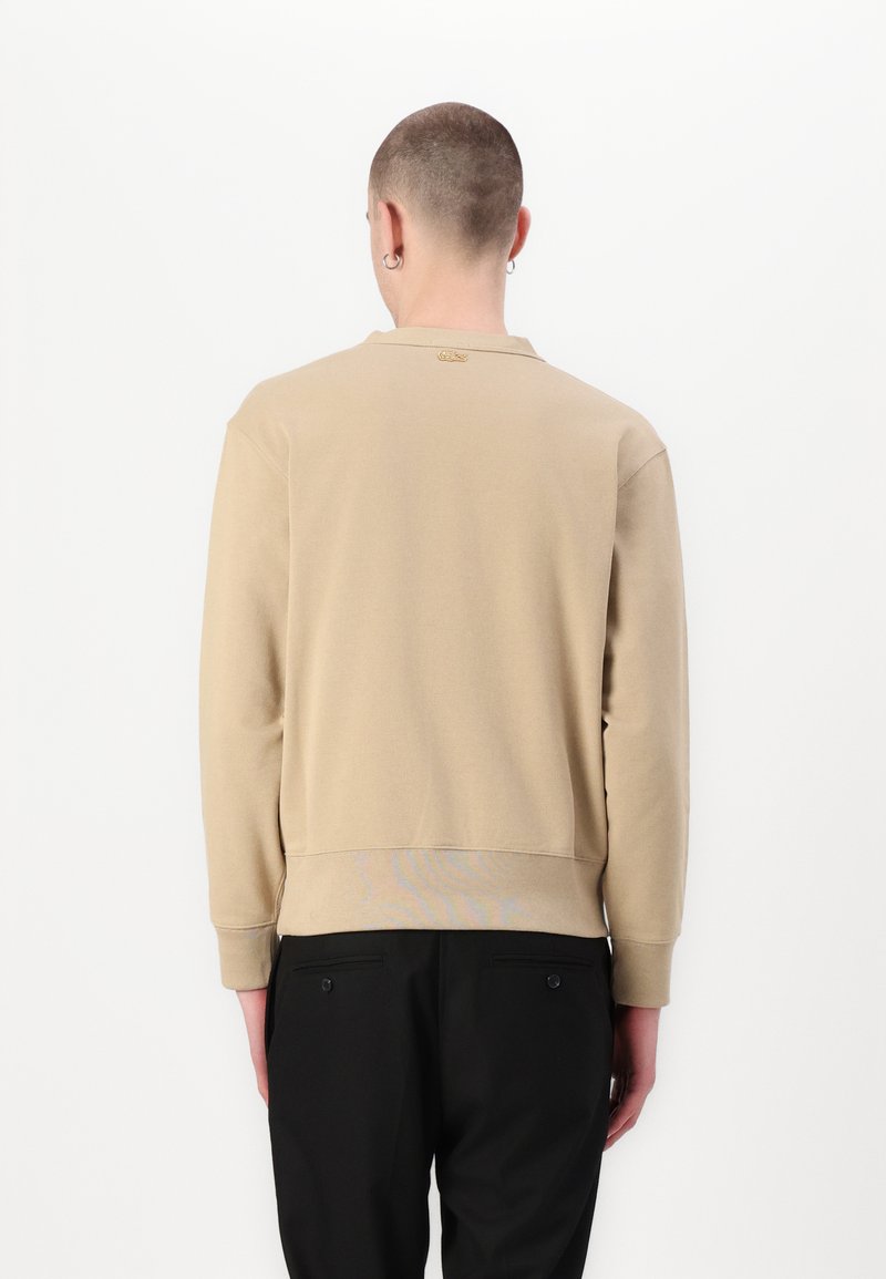 Sweatshirt beige à manches longues et col rond, avec une étiquette de marque discrète près du col. Porté avec un pantalon noir.
