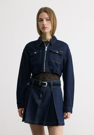 Młoda kobieta ubrana w ciemną, jeansową krótką kurtkę z klapowymi kieszeniami oraz plisowaną mini spódniczkę z czarnym paskiem na jednolitym tle.