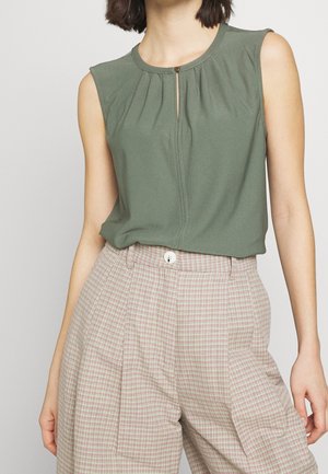 Femme portant un haut sans manches vert olive avec bouton œillet et pantalon à carreaux clair taille haute avec bouton avant et plis.