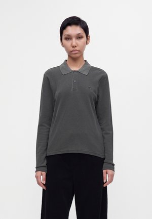 Giovane con capelli corti che indossa una polo a maniche lunghe grigio scuro e pantaloni neri, in piedi davanti a uno sfondo bianco semplice.