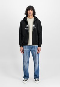 Crna zip-up dukserica s bijelim grafičkim logotipom, svijetlosmeđa majica ispod, uparena s svijetloplavim trapericama i crno-bijelim tenisicama.