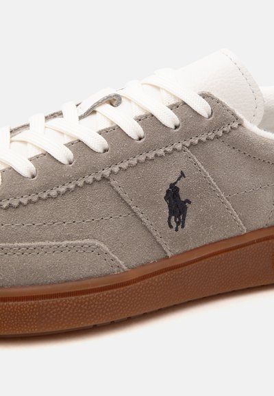 Close-up of beige suede sneaker with white laces, brown sole, and black polo player logo stitched on side.