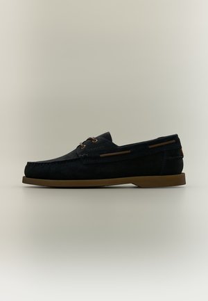 Chaussure bateau en daim noir avec lacets et semelle marron, présentée de profil sur un fond neutre.
