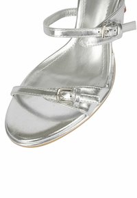 Sandalo con tacco in metallo argentato caratterizzato da sottili cinturini, un dettaglio con fibbia e una soletta in tessuto liscio. La suola è di colore marrone chiaro.