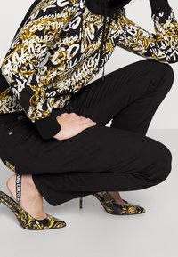 Mikina s kapucí s černým, bílým a zlatým vzorem, doplněná černými kalhotami a černými podpatky se zlatým květinovým vzorem a páskem s logem.