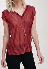 Top rojo de cuello en V con mangas cortas, que presenta un patrón de olas texturizado. Combinado con pantalones negros y un collar de péndulo plateado.