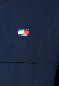 Tmavě modrá košile Tommy Jeans s nášivkou loga v červené, bílé a černé barvě. Hladká textura s kontrastním prošitím podél lemu.