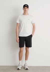 Tricou alb cu guler rotund și margine gri, împreună cu pantaloni scurți negri. Modelul poartă o șapcă neagră și adidași albi.