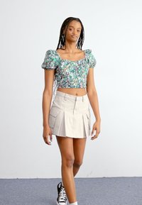 Virágmintás crop top puffos ujjakkal zöld és lila árnyalatokban, bézs pliszírozott szoknyával és fekete sportcipővel párosítva. Egyszerű, modern dizájn.