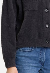 Cardigan grigio antracite morbido con una finitura testurizzata, dotato di una tasca frontale e chiusura a bottoni, abbinato a jeans in denim blu.