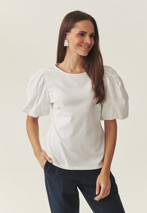 Woman with long brown hair wearing white puffed-sleeve blouse, silver square earrings, and dark pants, standing with one hand in pocket.