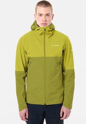 Junge Person trägt eine grüne Vaude Kapuzenjacke mit Frontreißverschluss und Reißverschlusstaschen an den Seiten, steht vor weißem Hintergrund.