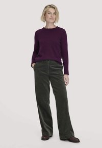 Lila Langarmpullover kombiniert mit dunkelgrünen Cord-Hose im weiten Schnitt. Das Modell steht mit den Händen in den Taschen und trägt braune Schuhe.