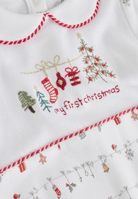 Vêtement blanc avec une broderie festive : "mon premier Noël", des chaussettes, des cadeaux et des motifs de fête, accentué de coutures rouges.