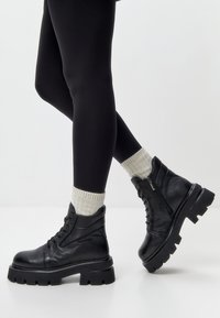Zwarte leren enkellaarsjes met dikke zolen, vetersluiting en rits aan de zijkant. Ribbelige grijze sokken contrasteren met strakke zwarte leggings.