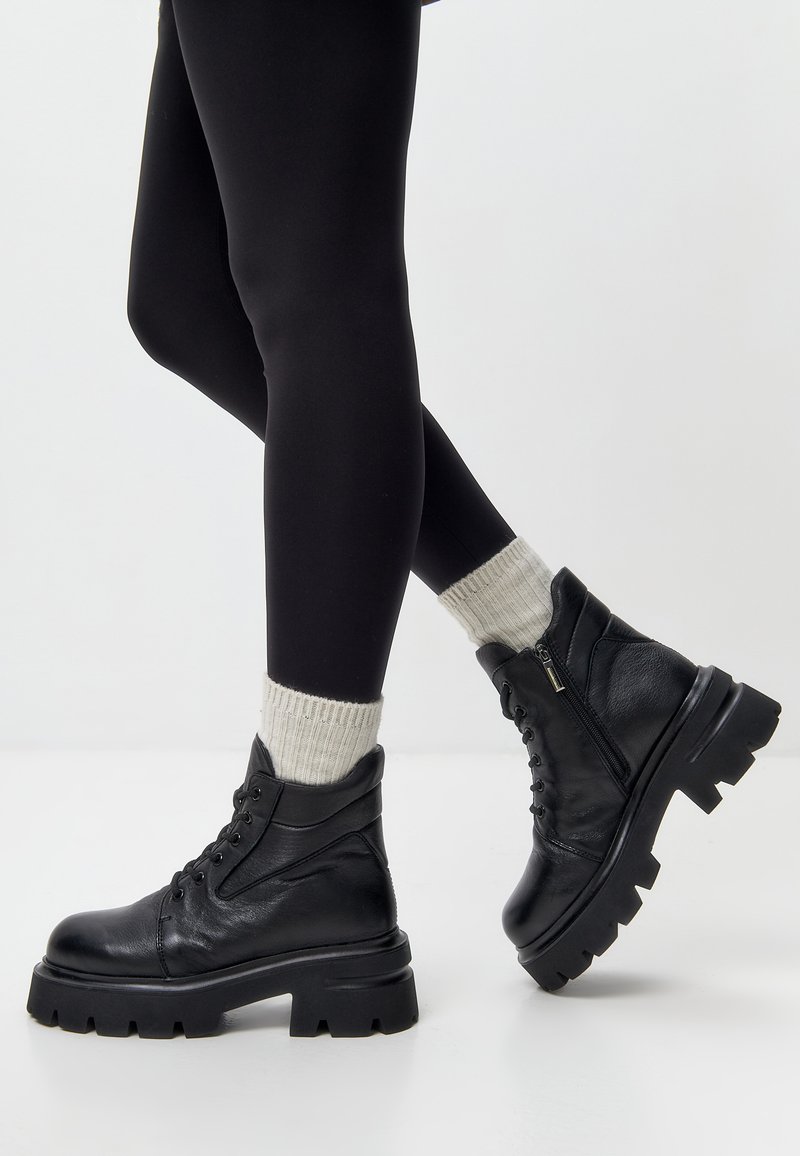 Zwarte leren enkellaarsjes met dikke zolen, vetersluiting en rits aan de zijkant. Ribbelige grijze sokken contrasteren met strakke zwarte leggings.