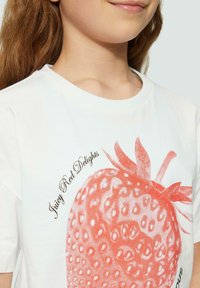 T-shirt en coton blanc avec un grand graphique de fraise orange affichant des graines texturées et le texte "Délices Rouges Juteux" en noir.