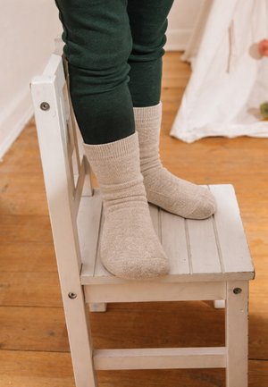 Child wearing beige socks and dark pants stands on a small white wooden chair on a wooden floor indoors.