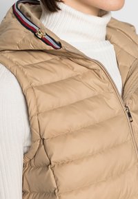 Beżowa kamizelka puffer z poziomymi przeszyciami, zapinana na zamek, z czerwono-niebieskim paskiem na szyi. Noszona na białym prążkowanym golfie.