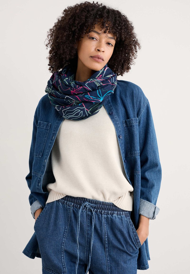 Cirkulær tørklæde i mørkeblå med farverige abstrakte bladmønstre. Båret over en cremefarvet sweater og en denimjakke, som har en afslappet pasform.